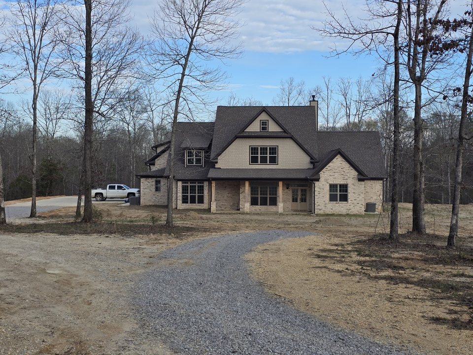 Front view of a home with light brick, a dark gabled roof, and timber porch posts in a wooded setting.