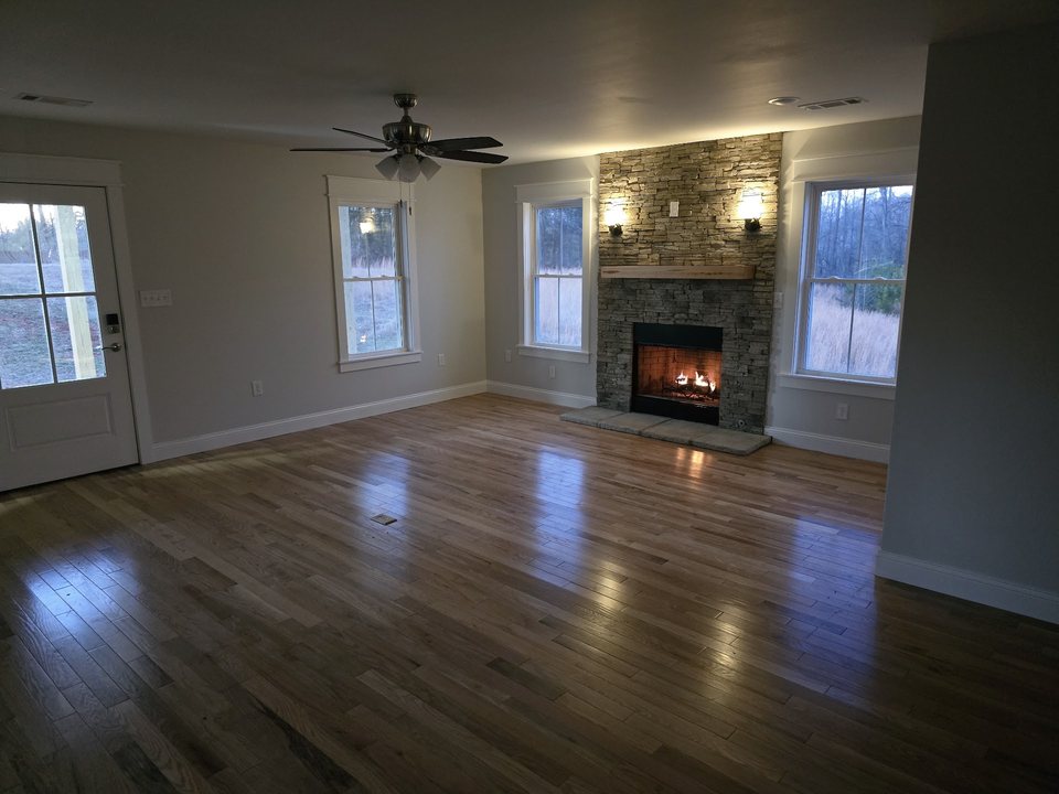 Living room with hardwood flooring and a lit stone fireplace between tall windows.